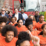 A group of men in suits are walking behind a group of teenagers in orange shirts