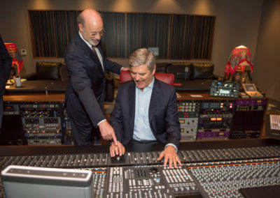 One man in a black suit is standing next to another man in a black suit sitting in front of a soundboard