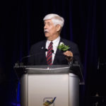 A man with white hair in a red tie and black suit is standing and speaking at a podium at the 2018 YCEA dinner