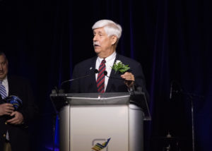 A man with white hair in a red tie and black suit is standing and speaking at a podium at the 2018 YCEA dinner