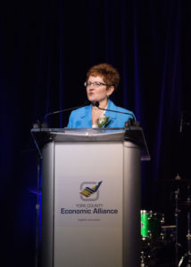 A woman with brown hair in a blue shirt is standing at a podium during the 2018 YCEA dinner