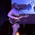 A man is standing on stage while singing into a microphone and playing a guitar at the 2018 YCEA dinner