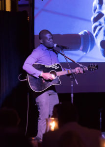 A man is standing on stage while singing into a microphone and playing a guitar at the 2018 YCEA dinner