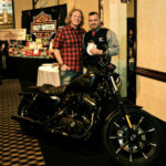 Two men are smiling and posing behind a black harley davidson motorcycle