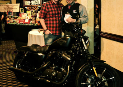Two men are smiling and posing behind a black harley davidson motorcycle