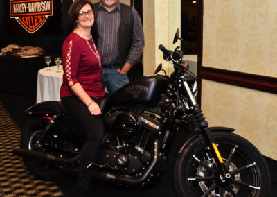 A man in a grey shirt and a woman in a red shirt are posing behind a black harley davidson motorcycle