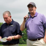 One man in a purple polo shirt is standing beside another man in a black polo shirt and is speaking into a microphone on a golf course