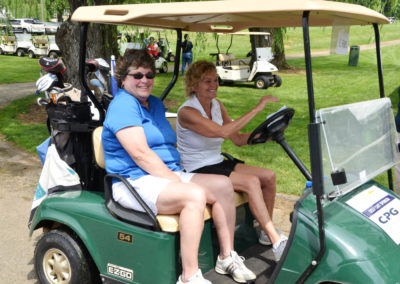 Two women wearing polo shirts and shorts are sitting in a golf cart on a golf course