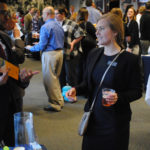 A woman in a black business suit is holding a cup of juice while speaking to a man in a black suit at a conference